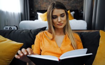 A woman with long hair is sitting on a couch, wearing an orange blouse, and reading a book. Behind her is a bed and yellow cushions. The room has a cozy, softly lit atmosphere.