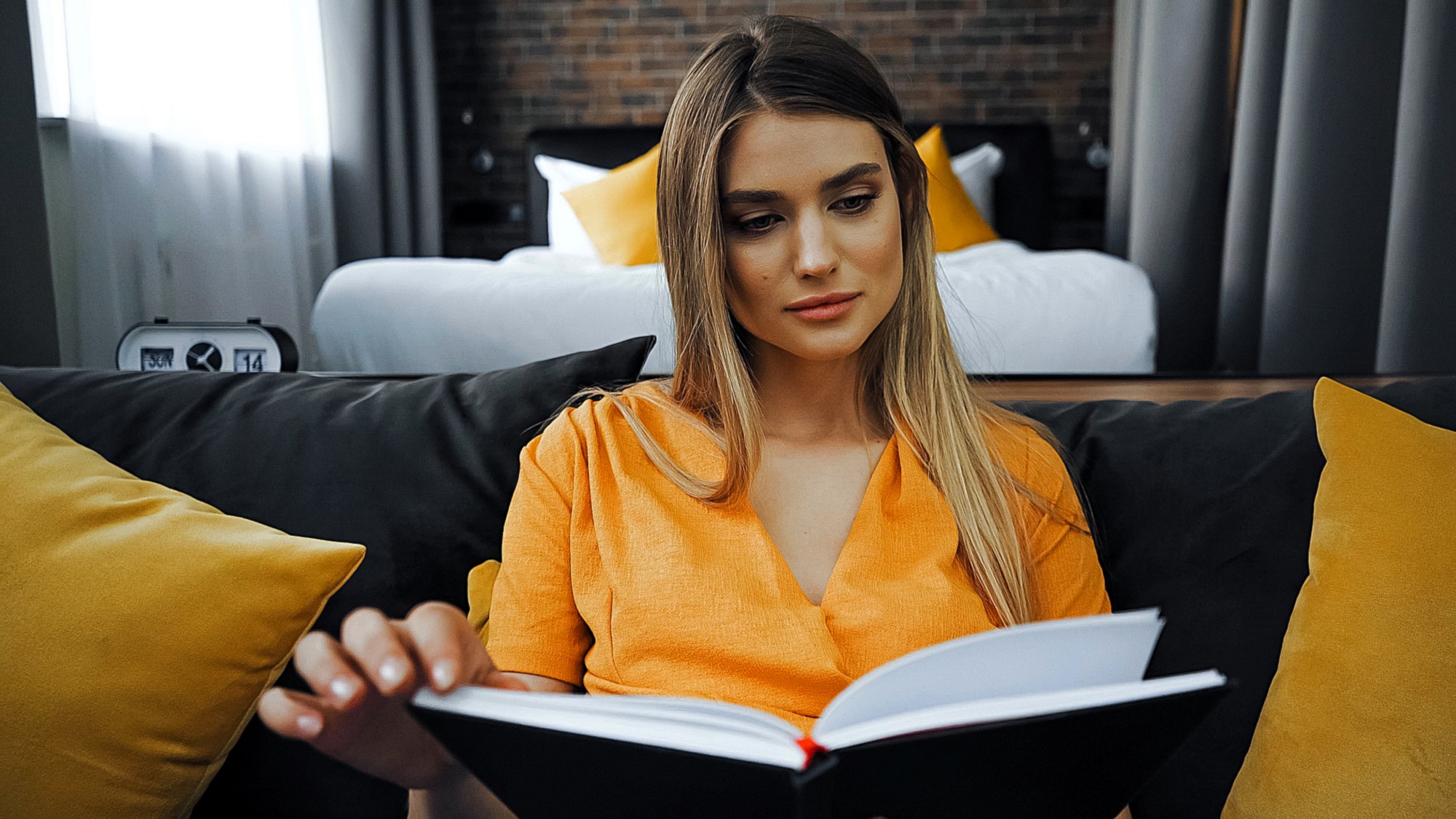 A woman with long hair is sitting on a couch, wearing an orange blouse, and reading a book. Behind her is a bed and yellow cushions. The room has a cozy, softly lit atmosphere.