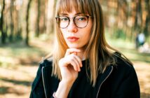 A person with long hair and glasses wearing a black jacket, standing in a forest. They have their hand under their chin and are looking directly at the camera with a thoughtful expression. The background is blurred with trees.