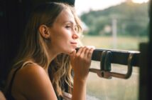 A woman with long blonde hair gazes thoughtfully out of a train window, resting her hands on the ledge. The blurred landscape outside suggests motion, with greenery visible in the background. The light is soft and natural, conveying a serene atmosphere.