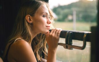 A woman with long blonde hair gazes thoughtfully out of a train window, resting her hands on the ledge. The blurred landscape outside suggests motion, with greenery visible in the background. The light is soft and natural, conveying a serene atmosphere.