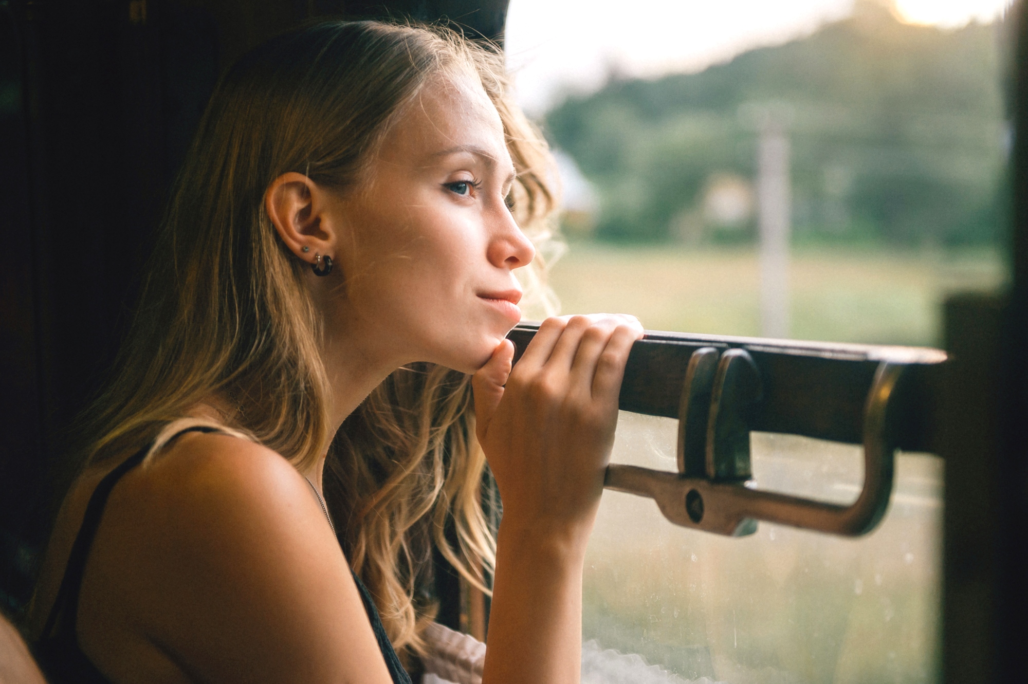 A woman with long blonde hair gazes thoughtfully out of a train window, resting her hands on the ledge. The blurred landscape outside suggests motion, with greenery visible in the background. The light is soft and natural, conveying a serene atmosphere.