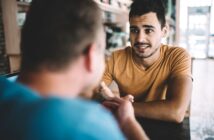 A man in an orange shirt sits at a table, engaged in a conversation with another person in a blue shirt. They are in a bright setting with shelves visible in the background. The man appears to be listening attentively.