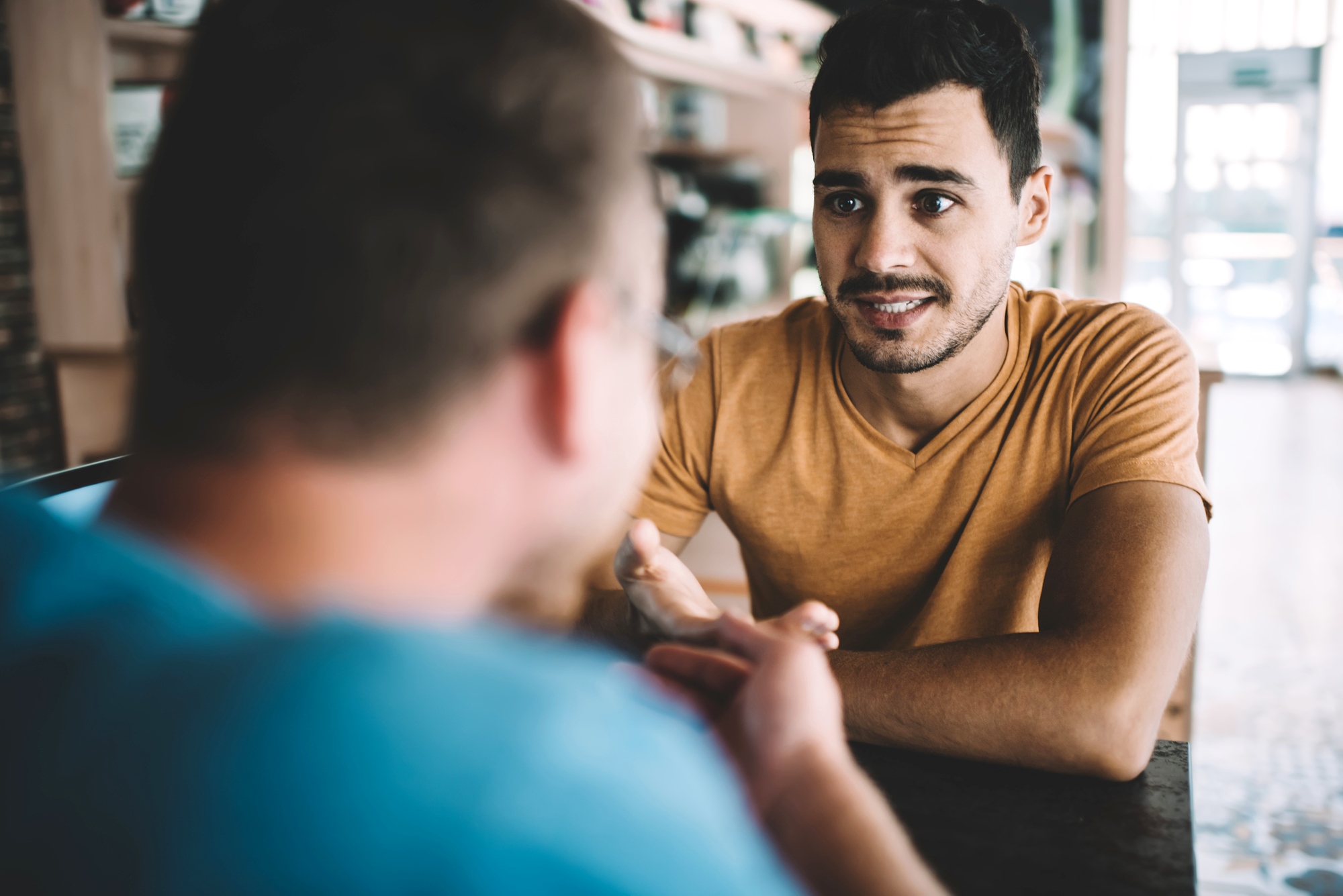 A man in an orange shirt sits at a table, engaged in a conversation with another person in a blue shirt. They are in a bright setting with shelves visible in the background. The man appears to be listening attentively.