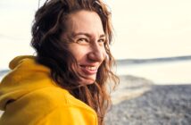 A woman with long brown hair smiles joyfully at the camera on a beach. She is wearing a yellow hoodie, and the background features a serene, blurred view of the ocean and shoreline.