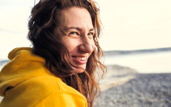 A woman with long brown hair smiles joyfully at the camera on a beach. She is wearing a yellow hoodie, and the background features a serene, blurred view of the ocean and shoreline.