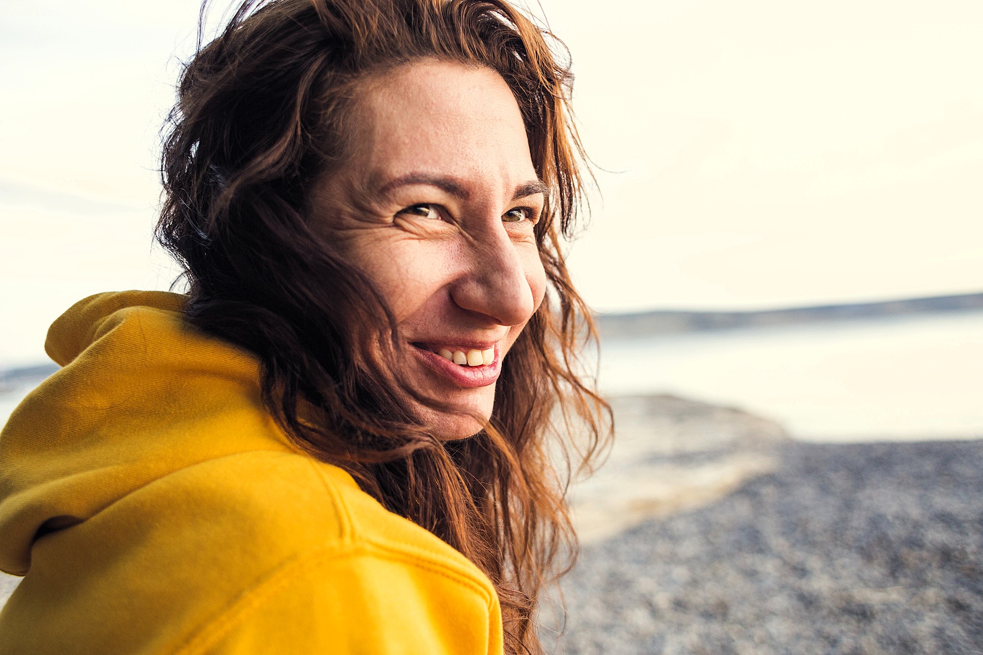 A woman with long brown hair smiles joyfully at the camera on a beach. She is wearing a yellow hoodie, and the background features a serene, blurred view of the ocean and shoreline.