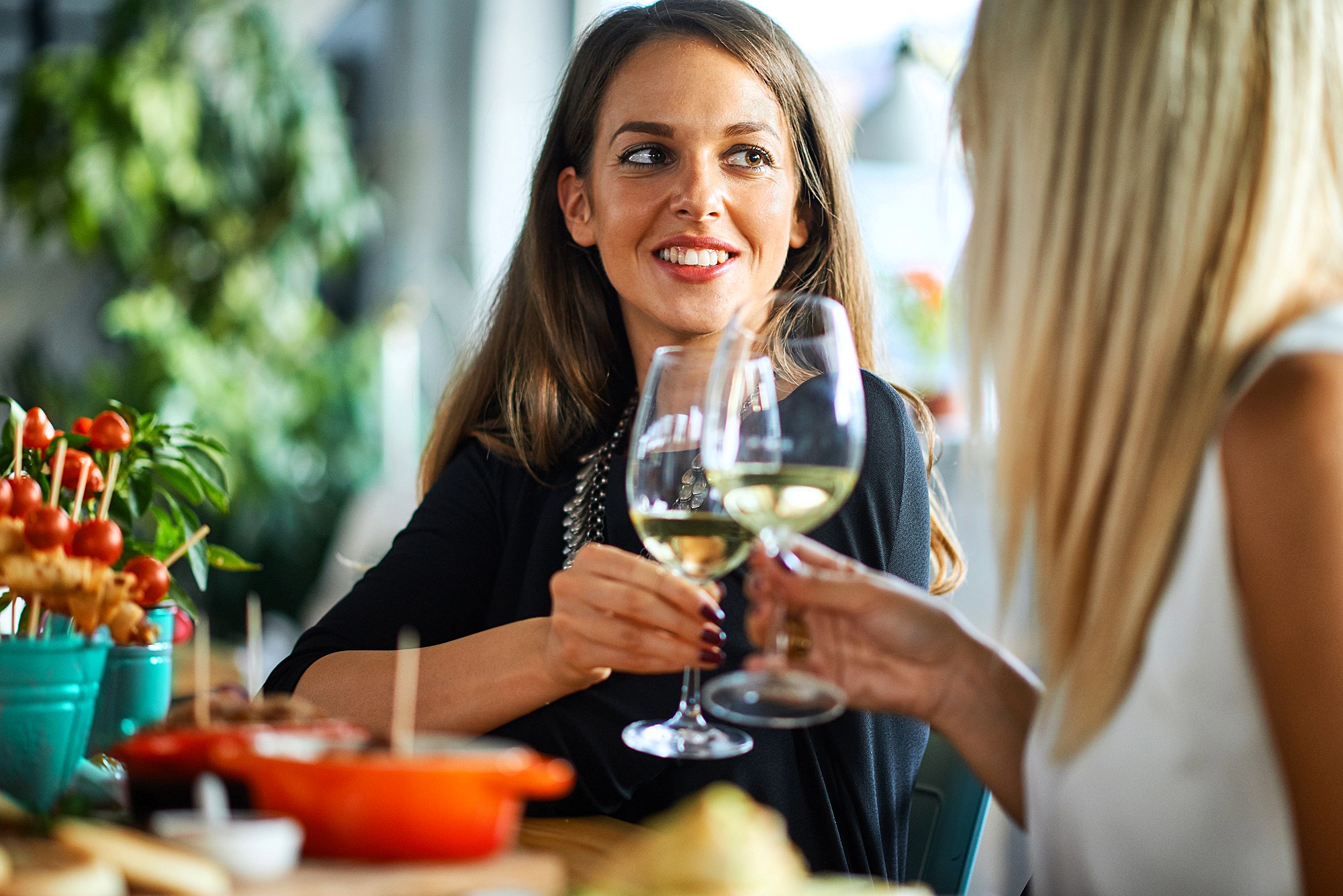 Two women smiling and clinking wine glasses at a dining table. The table is adorned with various dishes and colorful skewers. They appear to be enjoying a casual, cheerful meal in a lively setting.