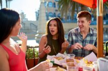 Three friends are sitting outdoors at a restaurant table, enjoying food and drinks. Two women and a man engage in lively conversation. The table is filled with plates of food, and there is a sunny atmosphere with palm trees in the background.