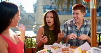 Three friends are sitting outdoors at a restaurant table, enjoying food and drinks. Two women and a man engage in lively conversation. The table is filled with plates of food, and there is a sunny atmosphere with palm trees in the background.