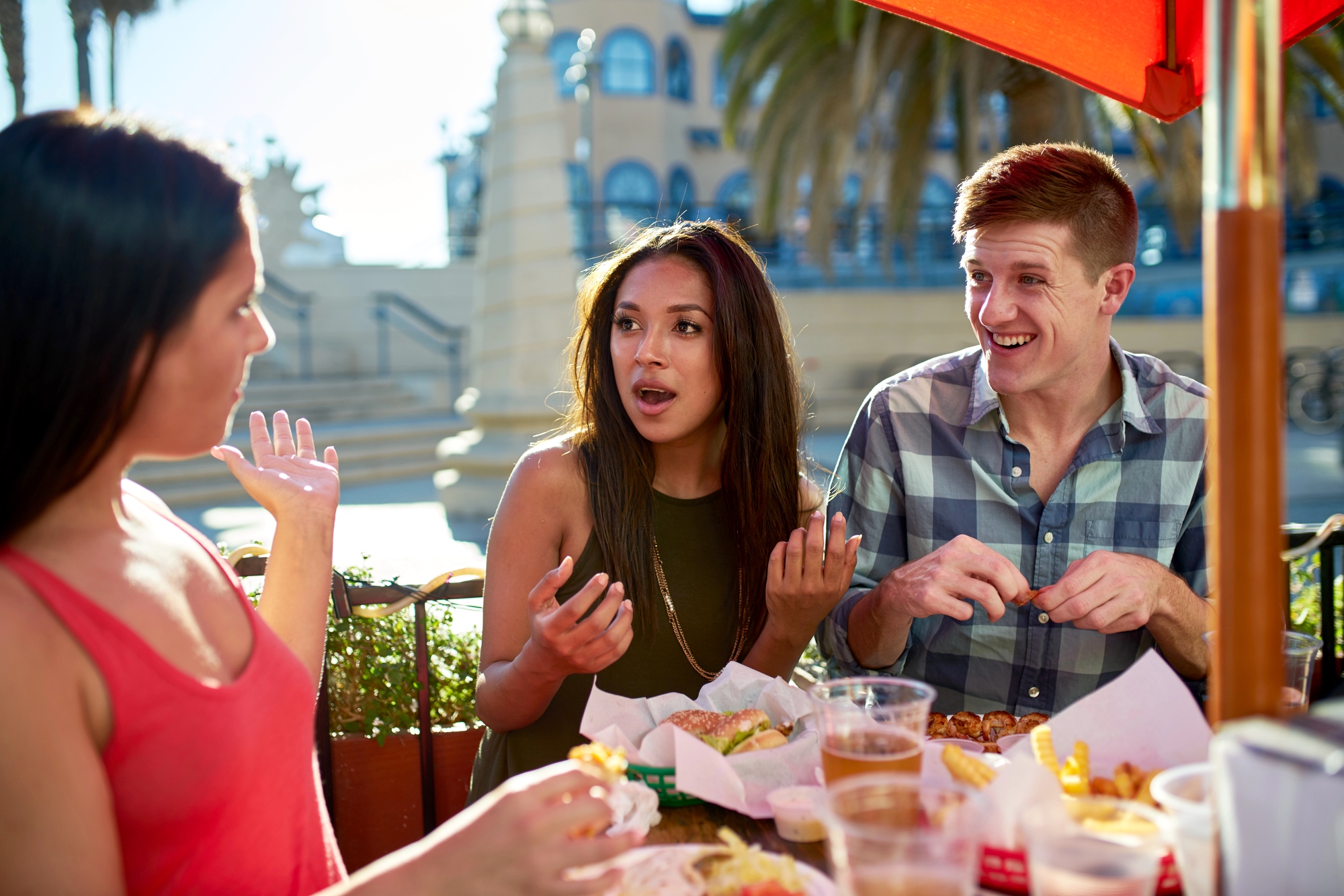 Three friends are sitting outdoors at a restaurant table, enjoying food and drinks. Two women and a man engage in lively conversation. The table is filled with plates of food, and there is a sunny atmosphere with palm trees in the background.