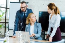 Three business professionals are having a discussion in an office. A woman sitting at a desk gestures while talking to two colleagues, a man and another woman, who are both standing. A laptop and water bottles are on the table.