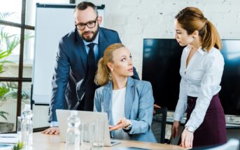 Three business professionals are having a discussion in an office. A woman sitting at a desk gestures while talking to two colleagues, a man and another woman, who are both standing. A laptop and water bottles are on the table.