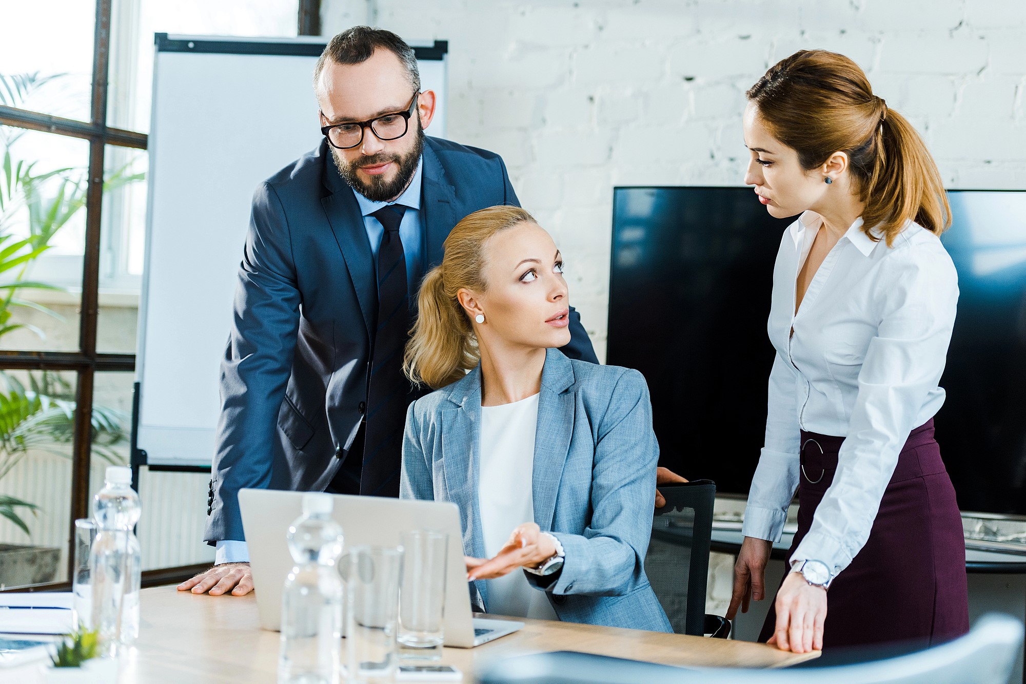 Three business professionals are having a discussion in an office. A woman sitting at a desk gestures while talking to two colleagues, a man and another woman, who are both standing. A laptop and water bottles are on the table.