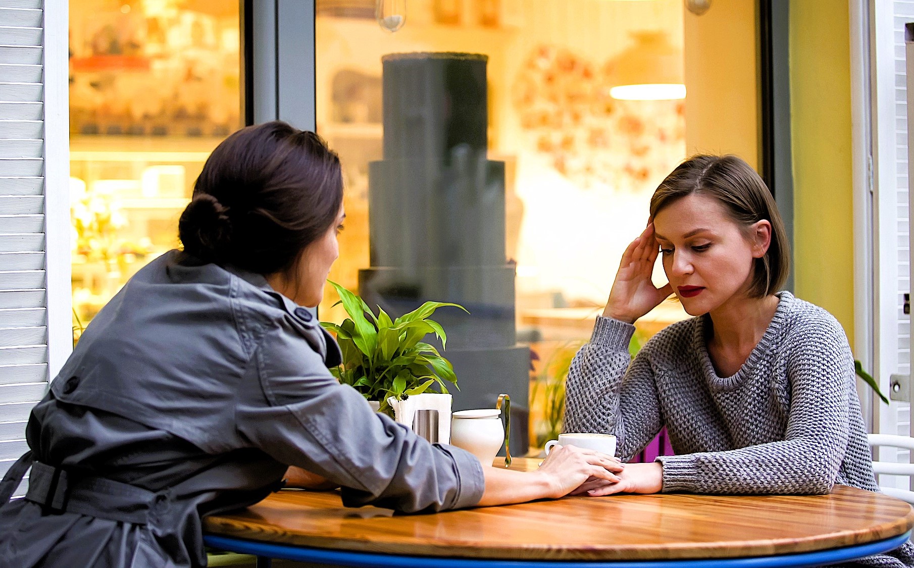 Two women sit across from each other at an outdoor café table. One woman looks concerned and holds a cup, while the other, in a gray coat, gently touches her hand. A potted plant is on the table, and a window displays a cake inside.