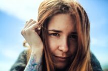 A woman with long red hair gazes into the camera, smiling slightly. She holds a hand near her face, revealing a tattoo on her forearm. The sky is clear and blue in the background.