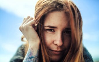 A woman with long red hair gazes into the camera, smiling slightly. She holds a hand near her face, revealing a tattoo on her forearm. The sky is clear and blue in the background.