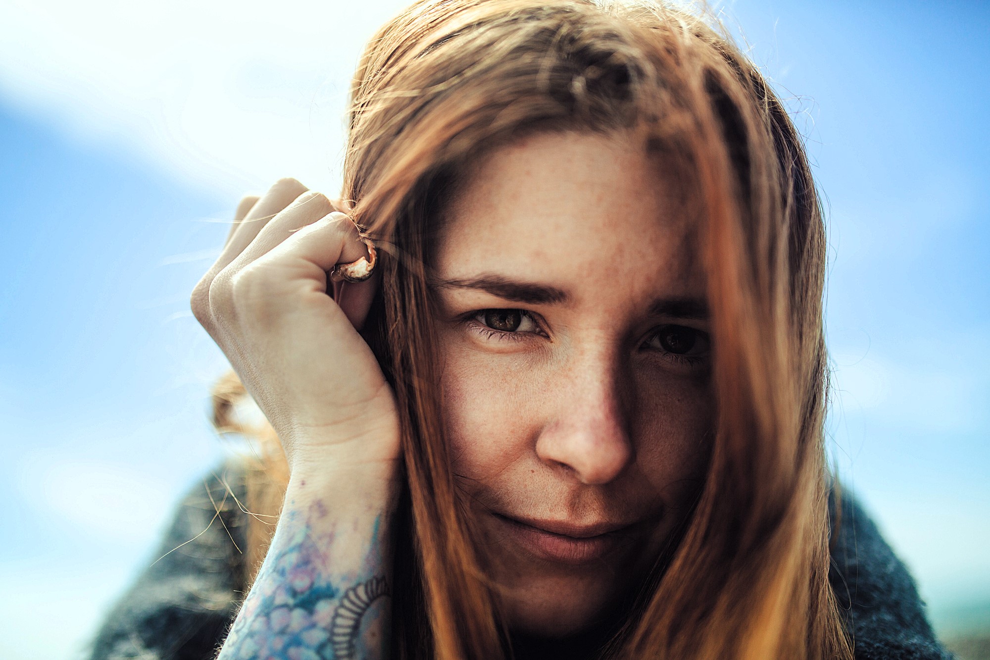 A woman with long red hair gazes into the camera, smiling slightly. She holds a hand near her face, revealing a tattoo on her forearm. The sky is clear and blue in the background.