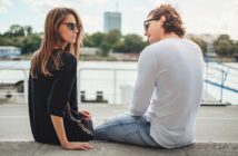 A woman and a man sit on a stone ledge by a river, both wearing sunglasses. The woman in a black shirt faces the camera, while the man in a white shirt looks at her. Trees and a tall building are visible across the water.