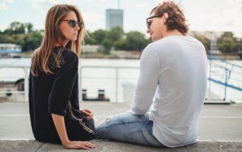 A woman and a man sit on a stone ledge by a river, both wearing sunglasses. The woman in a black shirt faces the camera, while the man in a white shirt looks at her. Trees and a tall building are visible across the water.