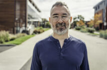 A man with a beard and glasses is smiling while standing on a quiet street. He is wearing a blue shirt. The background includes modern buildings and some trees with autumn foliage under a partly cloudy sky.