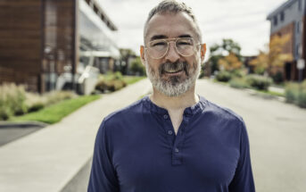 A man with a beard and glasses is smiling while standing on a quiet street. He is wearing a blue shirt. The background includes modern buildings and some trees with autumn foliage under a partly cloudy sky.