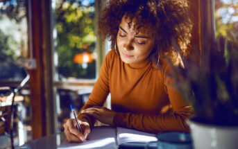 A person with curly hair, wearing a brown long-sleeve shirt, sits at a table writing in a notebook. The scene is warmly lit, with a window showing greenery outside, creating a cozy and focused atmosphere.