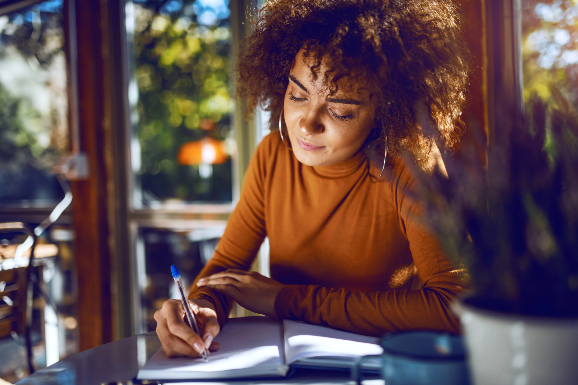 A person with curly hair, wearing a brown long-sleeve shirt, sits at a table writing in a notebook. The scene is warmly lit, with a window showing greenery outside, creating a cozy and focused atmosphere.