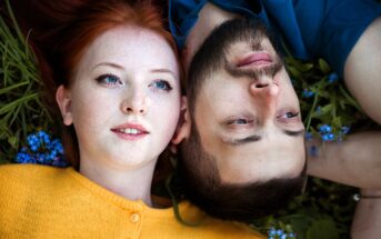 A red-haired woman in a yellow sweater and a bearded man in a blue shirt lie on grass, surrounded by small blue flowers. They are closely positioned, both gazing upwards.