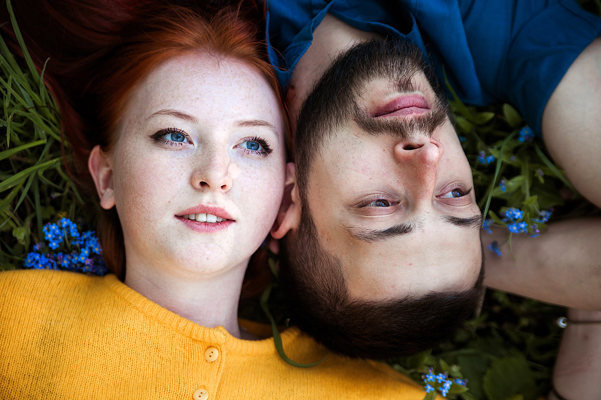 A red-haired woman in a yellow sweater and a bearded man in a blue shirt lie on grass, surrounded by small blue flowers. They are closely positioned, both gazing upwards.