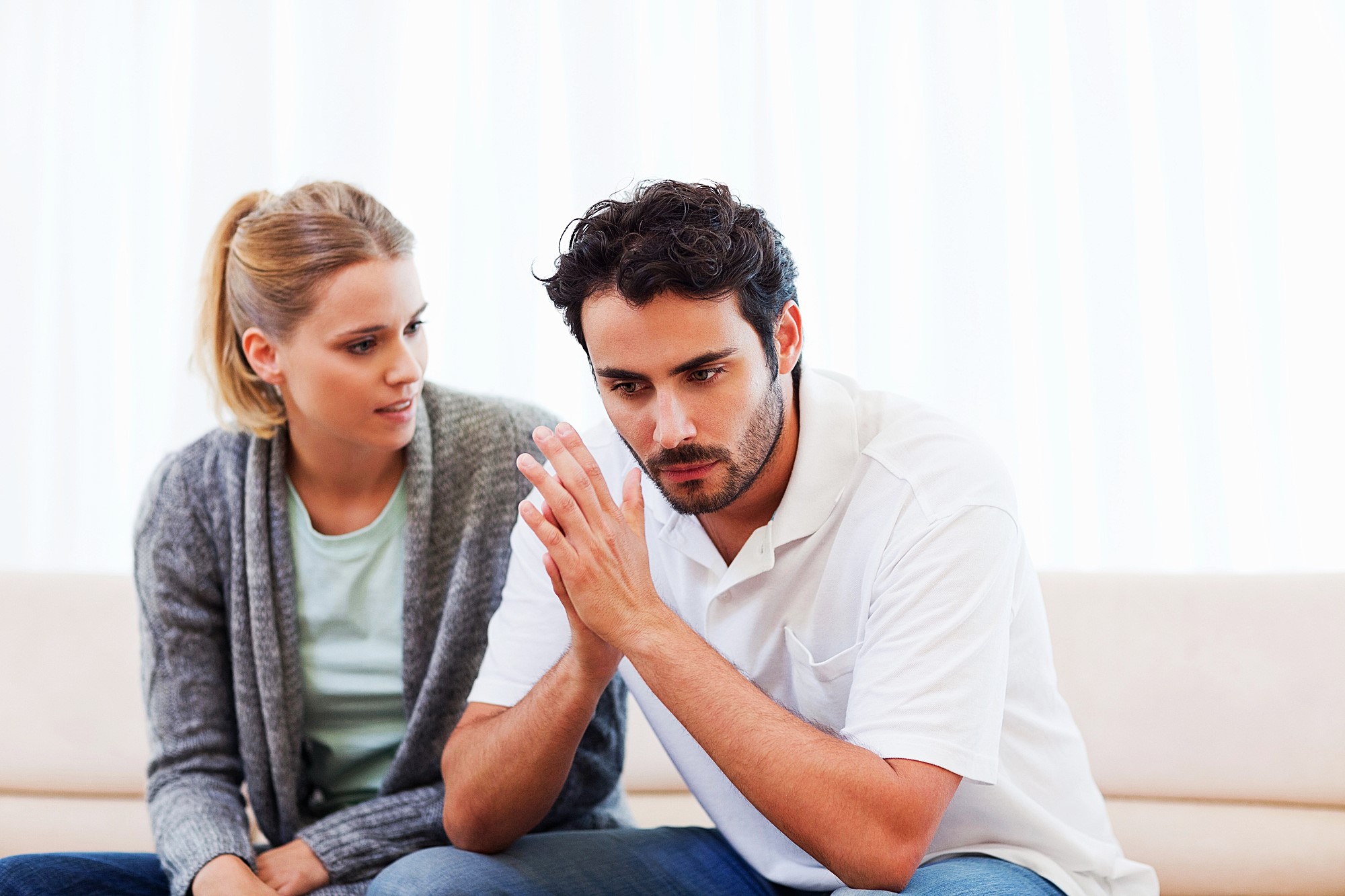 A woman sits next to a man on a couch. She appears concerned, looking at him while he gazes downward with his hands clasped together, seemingly deep in thought. The background is softly lit with white curtains.