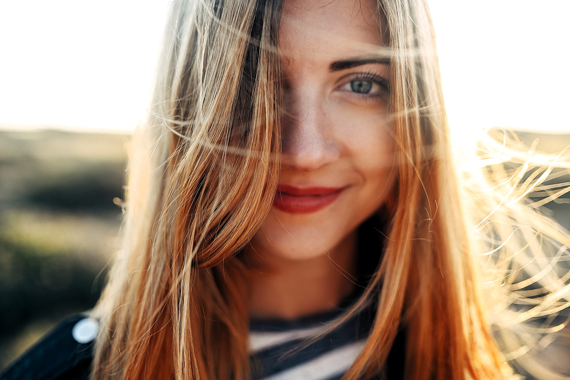 A close-up of a person with long hair smiling softly, captured outdoors in bright, natural light. Strands of hair are gently blowing across their face, creating a warm and serene atmosphere. The background is blurred, emphasizing the subject.