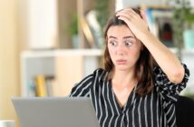 A woman with long brown hair wearing a black and white striped shirt looks surprised while holding her head with one hand and staring at a laptop screen. The background shows shelves with books and plants.