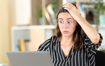 A woman with long brown hair wearing a black and white striped shirt looks surprised while holding her head with one hand and staring at a laptop screen. The background shows shelves with books and plants.