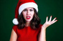 A woman wearing a Santa hat and a red top against a green background looks puzzled, with one hand raised. She has wavy brown hair and is wearing red lipstick and nail polish.