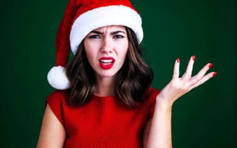 A woman wearing a Santa hat and a red top against a green background looks puzzled, with one hand raised. She has wavy brown hair and is wearing red lipstick and nail polish.