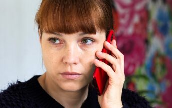 A woman with red hair is holding a red smartphone to her ear, looking to the side with a thoughtful expression. She is wearing a dark sweater, and the background is colorful and abstract.