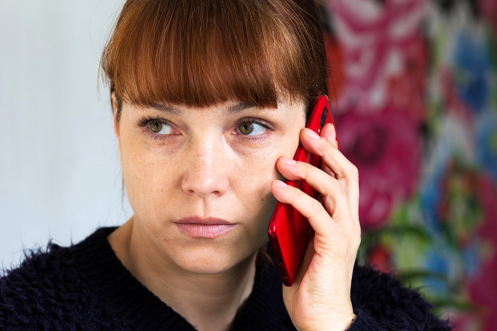 A woman with red hair is holding a red smartphone to her ear, looking to the side with a thoughtful expression. She is wearing a dark sweater, and the background is colorful and abstract.