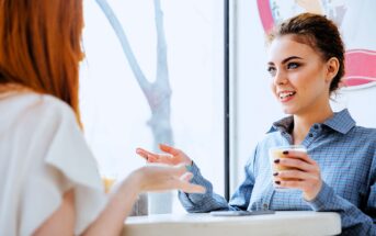 Two women sitting at a table in a café, engaged in conversation. One woman has curly hair and is holding a coffee cup, while the other, with long red hair, gestures with her hand. Natural light streams through the large window next to them.