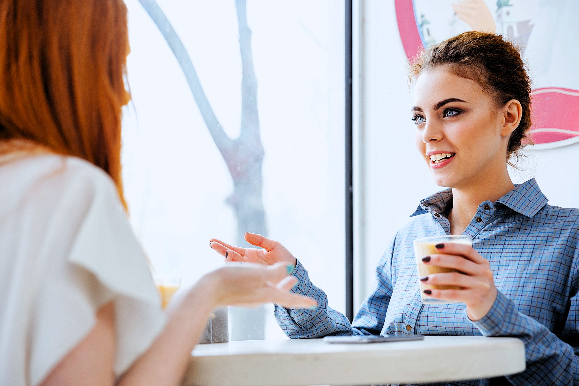 Two women sitting at a table in a café, engaged in conversation. One woman has curly hair and is holding a coffee cup, while the other, with long red hair, gestures with her hand. Natural light streams through the large window next to them.