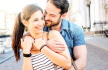 A couple smiles warmly at each other as the man hugs the woman from behind on a sunny street. She wears a striped top and he wears a denim shirt. They are standing near a historic building in an urban setting.