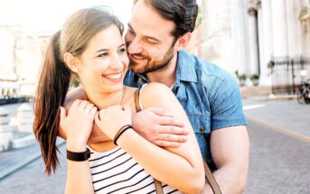 A couple smiles warmly at each other as the man hugs the woman from behind on a sunny street. She wears a striped top and he wears a denim shirt. They are standing near a historic building in an urban setting.