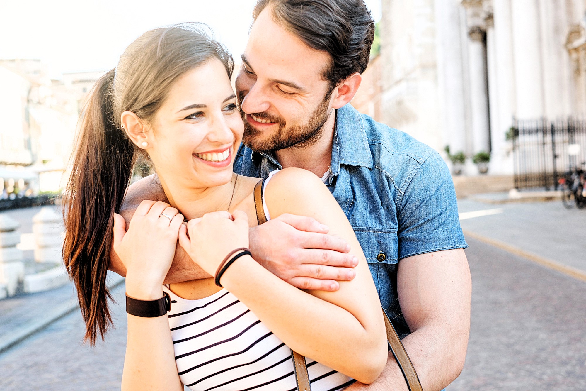 A couple smiles warmly at each other as the man hugs the woman from behind on a sunny street. She wears a striped top and he wears a denim shirt. They are standing near a historic building in an urban setting.