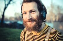 A smiling man with a full beard wearing headphones and a yellow and black striped shirt stands outdoors. The background is blurred, showing trees and soft sunlight.