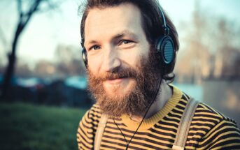 A smiling man with a full beard wearing headphones and a yellow and black striped shirt stands outdoors. The background is blurred, showing trees and soft sunlight.