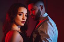 A woman with long dark hair in a black dress stands in the foreground, gazing confidently at the camera. A man with a beard and short hair, wearing a white shirt and tie, stands behind her. They are lit dramatically with red and blue lighting.