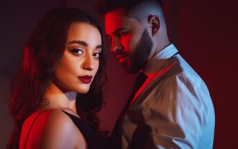 A woman with long dark hair in a black dress stands in the foreground, gazing confidently at the camera. A man with a beard and short hair, wearing a white shirt and tie, stands behind her. They are lit dramatically with red and blue lighting.