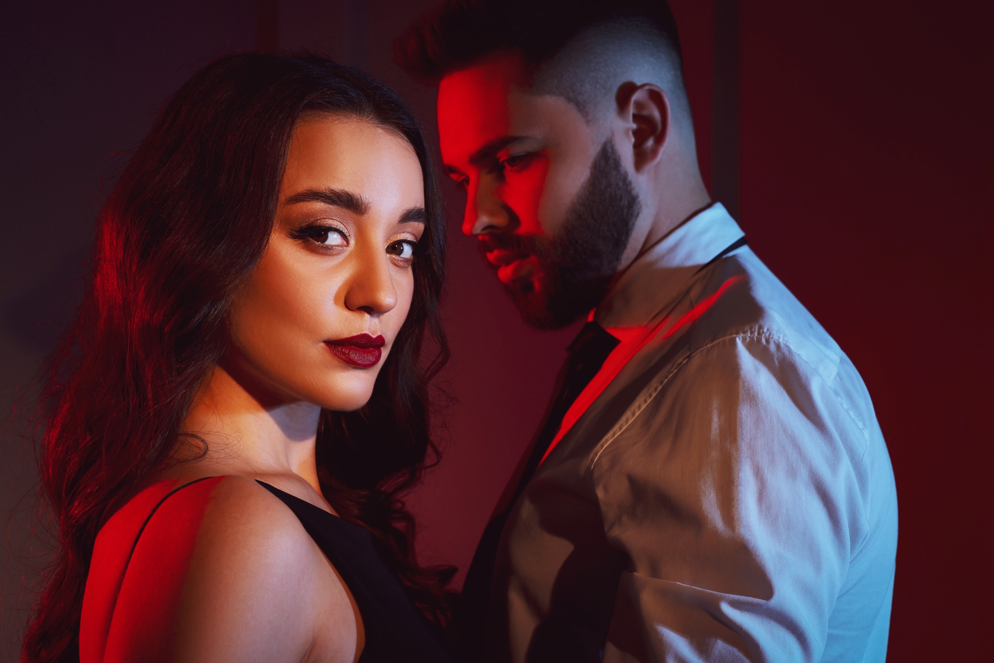 A woman with long dark hair in a black dress stands in the foreground, gazing confidently at the camera. A man with a beard and short hair, wearing a white shirt and tie, stands behind her. They are lit dramatically with red and blue lighting.