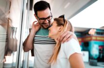 A man and woman are standing together outdoors, smiling and laughing. The man wears glasses and a striped shirt, gently wrapping his arm around the woman's shoulder. The woman, in a white shirt, looks up at him fondly. They are near a glass wall.