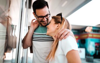 A man and woman are standing together outdoors, smiling and laughing. The man wears glasses and a striped shirt, gently wrapping his arm around the woman's shoulder. The woman, in a white shirt, looks up at him fondly. They are near a glass wall.